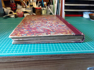 A massive book lying on a green cutting mat. It has maroon, gray, blue, and yellow marbled paper on the front.